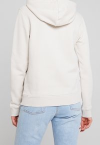 Vaaleanbeige huppari, jossa on nyörikapussi, resorit hihansuissa ja vyötäröllä. Pehmeä, teksturoitu kangas näytetään vaaleansinisien farkkujen kanssa.