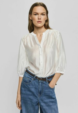 Kvinde iført en hvid knappet bluse med trekvartærmer og højttaljede blå jeans med et brunt bælte, stående med den ene hånd i lommen.