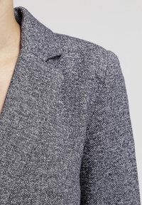 Grijze, gestructureerde blazer met een notch lapel en een subtiel patroon. De stof lijkt zacht en iets rekbaar. Close-up van het kraaggebied.