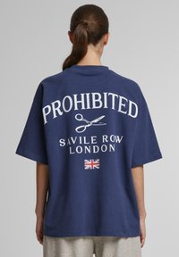 Mørkeblå oversized t-shirt med hvid "FORBUDT" tekst, saks grafik og "SAVILE ROW LONDON" nedenfor; har et britisk flag som accent.