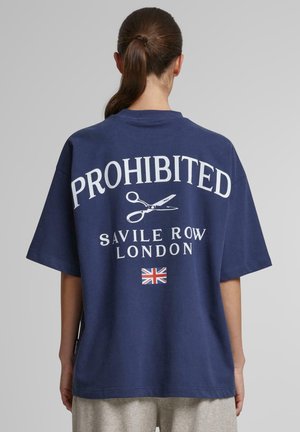 Mørkeblå oversized t-shirt med hvid "FORBUDT" tekst, saks grafik og "SAVILE ROW LONDON" nedenfor; har et britisk flag som accent.