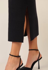 Zwarte geribbelde gebreide jurk met zijsplitten, gecombineerd met zwarte sandalen met hoge hakken en enkelbandjes met dunne hakken.