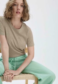 Khakifarbenes kurzärmliges T-Shirt aus weichem Stoff, kombiniert mit hellminzgrünen, mit Kordelzug versehenen Hosen. Modell sitzt auf einem Holzstuhl.