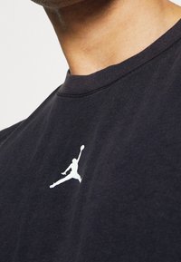 T-shirt en coton noir avec un logo Jordan blanc bien visible. Le col est côtelé et le tissu semble doux avec une surface légèrement texturée.