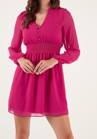 Fuchsia klänning med långa genomskinliga ärmar, V-ringning och smockad midja. Har knappar nedtill fram och en svävande kjol.