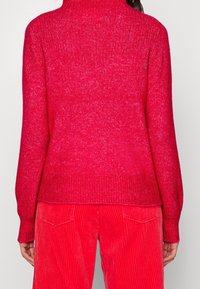 Maglione rosso a maglia con collo alto, finitura texturizzata e polsini a coste, abbinato a pantaloni in velluto a coste rossi con righe verticali.