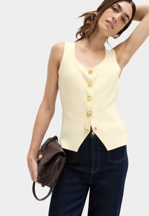 Mujer vistiendo una blusa sin mangas de color crema con cinco botones decorativos, jeans azul oscuro y sosteniendo un bolso marrón oscuro en una mano.