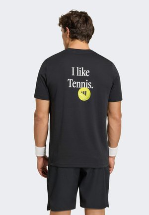 Mand iført sort tennisoutfit med teksten "Jeg kan lide tennis." og gul tennisbold grafik på bagsiden af skjorten, står med fronten væk fra os.