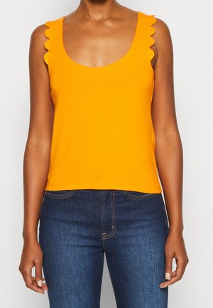 Kobieta ubrana w jasnopomarańczowy, bezrękawnik z falistymi krawędziami na ramionach i ciemnoniebieskie dżinsy na jednolitym tle.