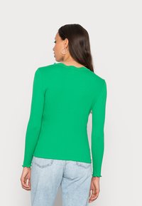 Top verde de manga longa com decote em cascata, design ajustado e tecido texturizado. Combinado com jeans de lavagem clara.