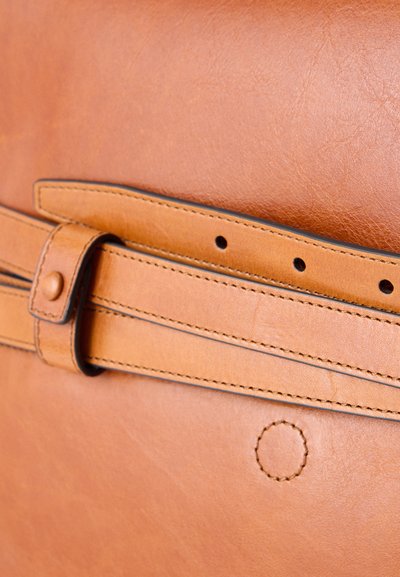 Gros plan sur une ceinture en cuir beige passée dans une lanière en cuir cousue avec des surpiqûres visibles et trois trous pour la ceinture.