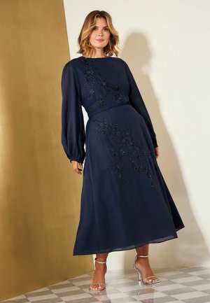 Frau trägt ein marineblaues, langärmeliges Midikleid mit floraler Stickerei und silberne Sandalen mit hohen Absätzen, steht auf einem Schachbrettboden.