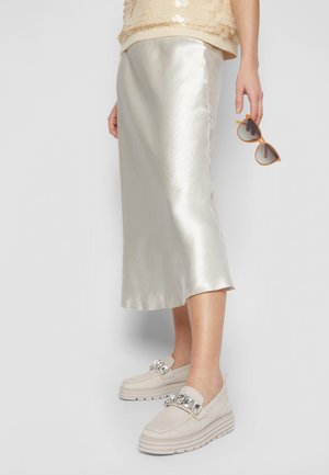 Kvinde iført en skinnende sølv midi-nederdel, prydede beige loafers og holder brune solbriller med ramme i hånden mod en hvid baggrund.