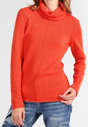 Femme portant un pull à col roulé orange vif à manches longues et un jean bleu déchiré, une main posée sur la hanche.