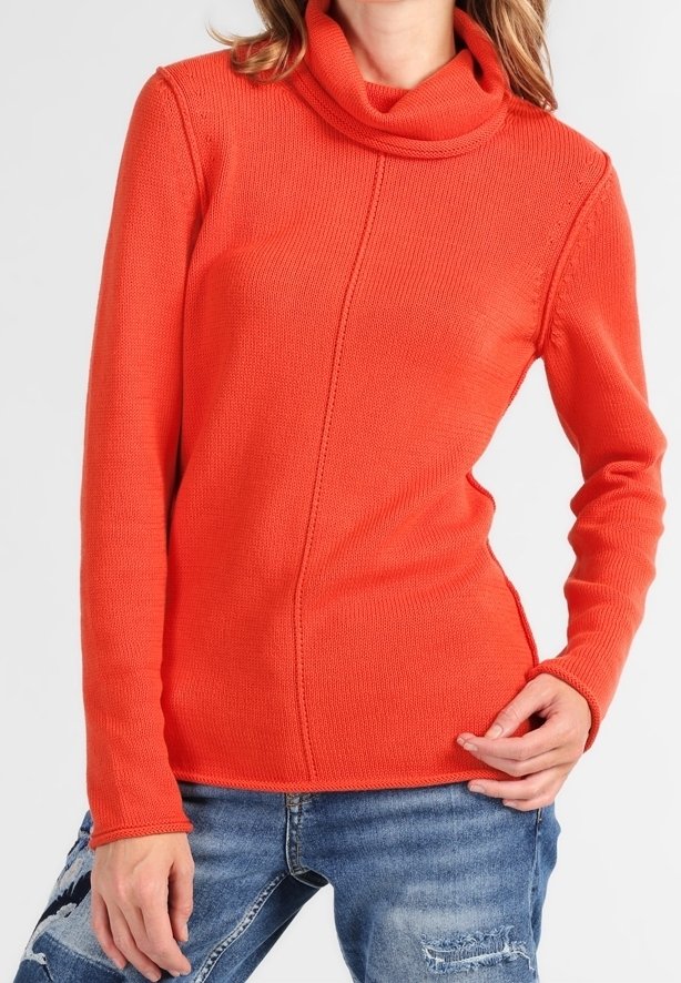 Femme portant un pull à col roulé orange vif à manches longues et un jean bleu déchiré, une main posée sur la hanche.