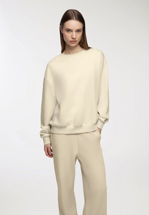 Felpa oversize color crema con scollatura rotonda, polsini a coste e fit rilassato, abbinata a pantaloni beige chiaro coordinati.
