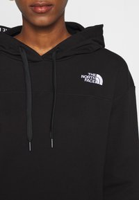 Gros plan d'une personne portant un sweat à capuche noir avec une poche avant et un logo blanc "The North Face" sur la poitrine gauche.