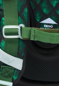 Zelený vzorovaný popruh batohu s plastovou sponou, vyrobený z polstrovaného černého síťového materiálu a s bílými logovými akcenty. Viditelné ergonomické designové prvky.