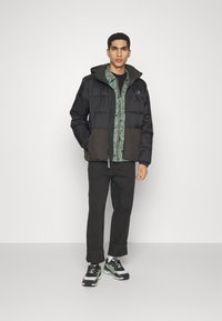 Black and dark green puffer jacket with a hood, featuring a geometric pattern on the inner lining, paired with black pants and sneakers.