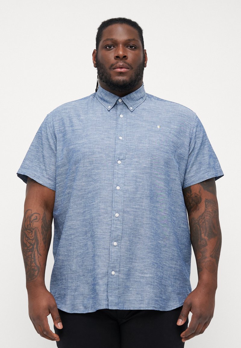 Camisa de botones de manga corta en una tela de chambray azul claro, con cuello, botones frontales y un pequeño logo en el pecho.