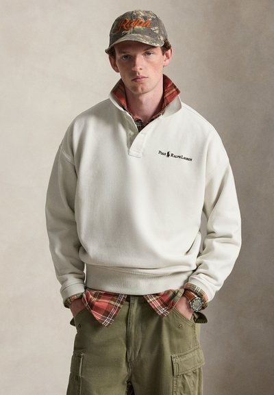 Νέος άνδρας φορώντας λευκό φούτερ Polo Ralph Lauren, κόκκινο καρό πουκάμισο από κάτω, χακί παντελόνι τύπου cargo, καμουφλάζ καπέλο μπέιζμπολ και ρολόι χειρός.