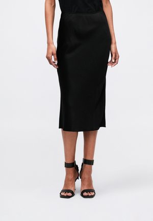 Woman wearing a black knee-length skirt with a side slit and black ankle-strap heeled sandals, standing on a plain white background.