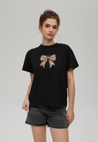 Camiseta de algodón negra con un gráfico de un lazo de estampado de leopardo en la parte frontal. El diseño es sencillo, con mangas cortas, cuello redondo y corte relajado.