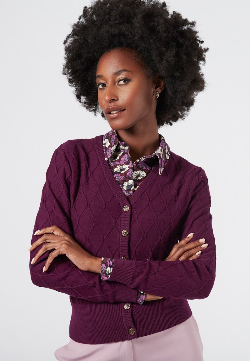 Mörklila stickad kofta med diamantmönster, v-ringad och femknappsstängning. Blommig skjorta med krage synlig under.