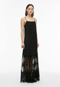 Femme portant une robe noire sans manches à fines bretelles, avec une superposition en dentelle florale transparente s'étendant jusqu'au sol, debout devant un fond blanc.
