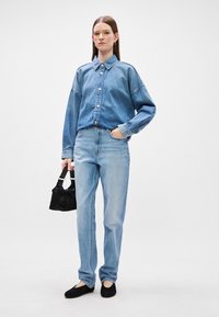 Denim jakna z gumbom na sprednji strani, dolgimi rokavi in klasičnim ovratnikom, v kombinaciji s svetlimi ravnimi kavbojkami in črnimi balerinkami.