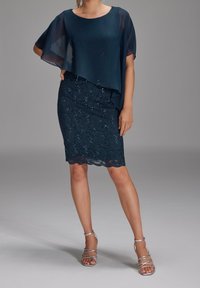 Femme portant une jupe en dentelle bleu foncé avec des sequins et un haut ample et transparent bleu foncé, associée à des sandales argentées à talons hauts et à brides.