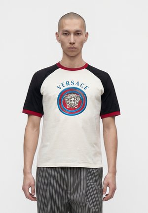 Homme portant un t-shirt à manches courtes avec des manches noires, une bordure rouge et un logo Versace avec Méduse dans des cercles bleus et rouges.