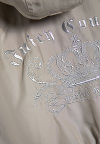 Бежова тъкан със сребриста бродирана текст "Juicy Couture" и детайлен дизайн на корона с декоративни завъртулки.