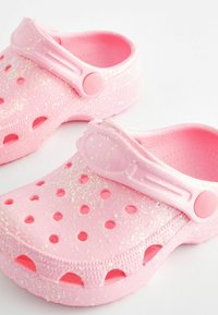 Rosa Clogs aus Kunststoff mit glänzender Oberfläche, ausgestattet mit Löchern zur Belüftung und einem Riemen für einen sicheren Sitz. Funkelnde Akzente überall.