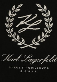 Melns audums ar baltu izšūtu lauvas vainagu un kursīva tekstu: "Karl Lagerfeld 21 RUE ST-GUILLAUME PARIS" biezā fontā.