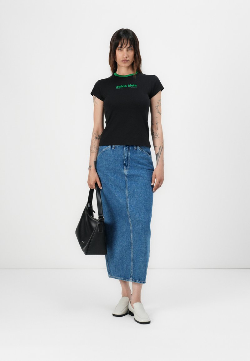 Schwarzes, figurbetontes T-Shirt mit grünem Logo, kombiniert mit einem langen blauen Jeansrock. Die Accessoires umfassen eine schwarze Schultertasche und weiße Slipper.