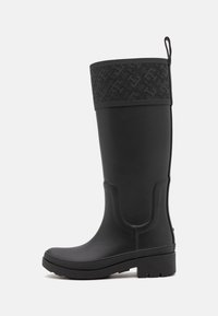 Botte de pluie en caoutchouc noir avec un bandeau supérieur noir à motifs, texture lisse, forme légèrement courbée et semelle plateforme basse texturée.