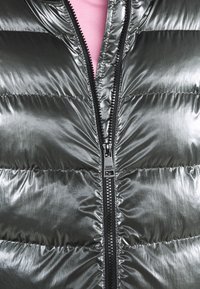 Gros plan sur une doudoune argentée brillante partiellement zippée, montrant une fermeture éclair noire et un aperçu d'une chemise rose en dessous.