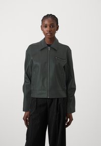 Tamsiai žalia odinė striukė su užtrauktuku, smailiu apykakle, priekinėmis užtrauktukinėmis kišenėmis ir lygiu paviršiumi. Derinama su juodai dryžuotomis kelnėmis.