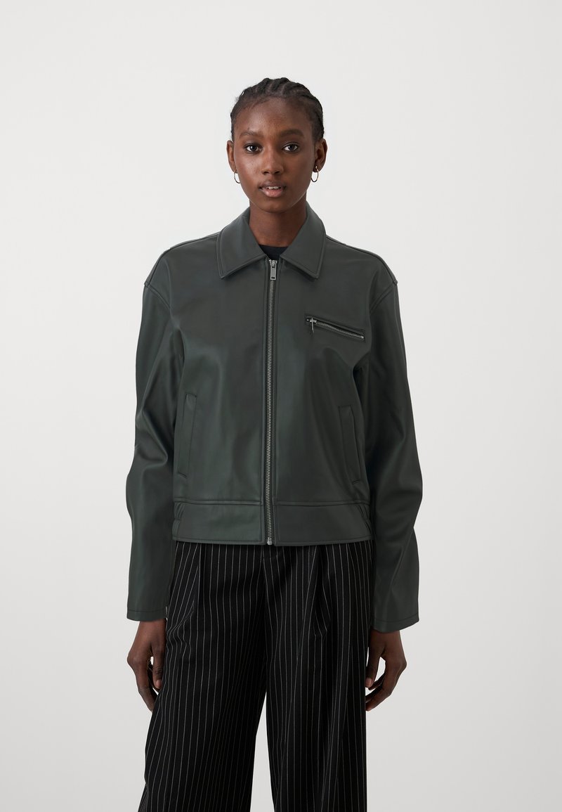 Tamsiai žalia odinė striukė su užtrauktuku, smailiu apykakle, priekinėmis užtrauktukinėmis kišenėmis ir lygiu paviršiumi. Derinama su juodai dryžuotomis kelnėmis.