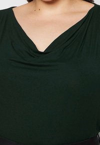 Skogsgrön topp med draperad, cowl-halsringning. Tillverkad av ett mjukt tyg, har den en figurnära silhuett och en subtil glans.