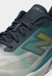 Κοντινή λήψη παπουτσιού τρεξίματος σε χρώμα τιρκουάζ και σκούρο μπλε, με σκούρες κορδόνια, υφασμάτινη υφή και στιλιζαρισμένο λογότυπο "N" στο πλάι.