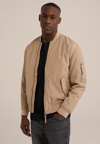 Béžová bomber bunda se zapínáním na zip, žebrovanými manžetami a bočními kapsami. Obsahuje kapsu na zip na levém rukávu. Stylizováno přes černé tričko.