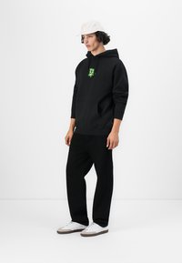 Sort hættetrøje med grønt grafisk logo, kengurulomme; parret med sorte bukser og hvide sneakers, akcentueret af en hvid kasket.