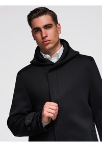 Jeune homme portant un manteau noir avec un col haut sur une chemise blanche, tenant l'avant du manteau près de la poitrine, fond neutre.