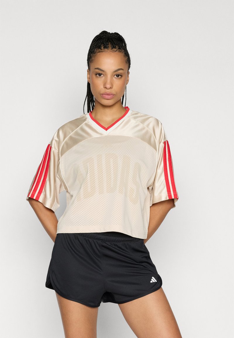 Top deportivo de malla beige con detalles en rojo, con un corte amplio y mangas cortas. Combinado con pantalones cortos deportivos negros y detalles de logotipo distintivos.
