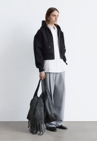 Schwarzer kurz geschnittener Hoodie mit Reißverschluss und zwei Fronttaschen, kombiniert mit einem weißen Hemd und grauen Hosen mit weitem Bein. Graue Wollhandtasche.