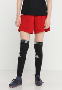 Pantalones cortos deportivos rojos con calcetines negros hasta la rodilla que tienen rayas blancas, combinados con botas de fútbol oscuras. El material parece transpirable.