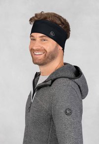 Zwarte stretchy hoofdband getoond op een glimlachende man, gedragen met een grijze rits-open hoodie. De hoofdband heeft een subtiel logo. Gladde textuur.