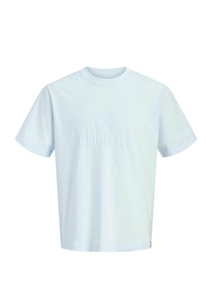 Teenage dreng står, iført en blå ternet skjorte over en hvid T-shirt, mørkeblå vide jeans og beige sko, smilende til kameraet.